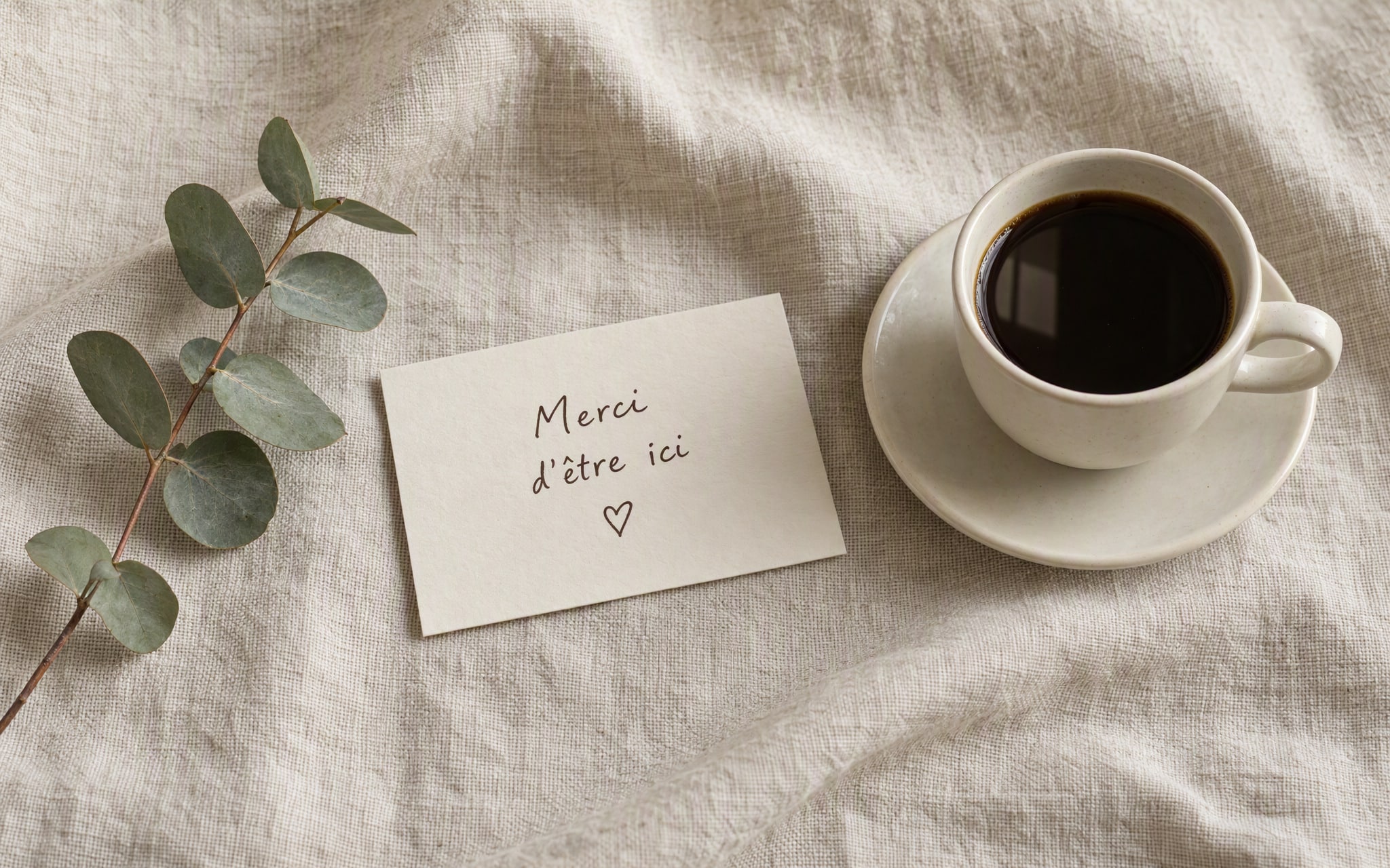 Une tasse de café et une carte Merci d’être ici sur du lin écru, accompagnées d’eucalyptus.